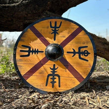 Viking round shield with golden birch wood, burgundy X-cross pattern, four Norse rune symbols, and black steel boss on outdoor wood chips