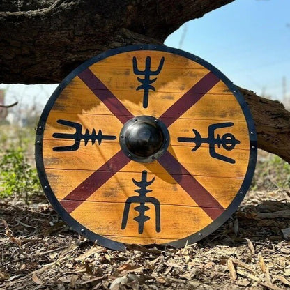 Viking round shield with golden birch wood, burgundy X-cross pattern, four Norse rune symbols, and black steel boss on outdoor wood chips