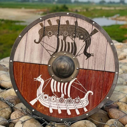 Viking longship round shield in dual-tone wood with dark and white drakkar design, metal rim with rivets, and bronze boss displayed on rocks outdoors