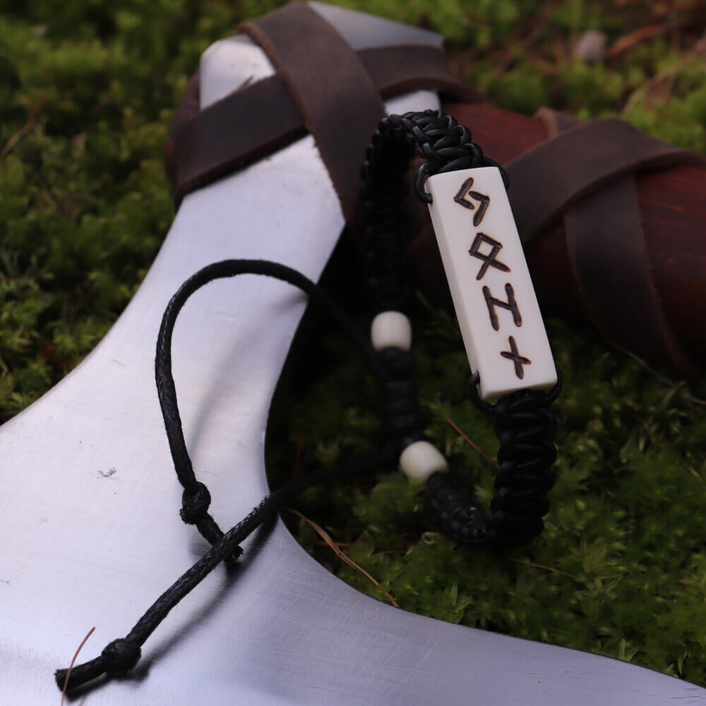 Black cord bracelet with white deer bone plate carved with Viking runes, resting on an axe blade outdoors.
