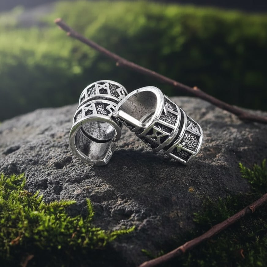 Two silver Viking Rune Spiral Wrap Beard Beads with engraved runes resting on a mossy stone outdoors.